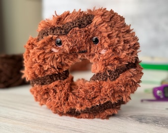 Könnte beinhalten: Ein braunes und schokoladenfarbenes Plüsch-Donut-Spielzeug mit einem niedlichen Gesicht. Der Donut hat einen braunen Guss und ein Schokoladenstreusel-Design.
