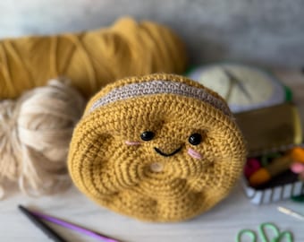 Könnte beinhalten: Ein handgefertigtes Häkelobjekt in Form einer lächelnden Kartoffel mit schwarzen Augen und einem gestickten Mund. Die Kartoffel ist senfgelb mit einem beigen Streifen. Häkelnadeln und Garn sind im Hintergrund.