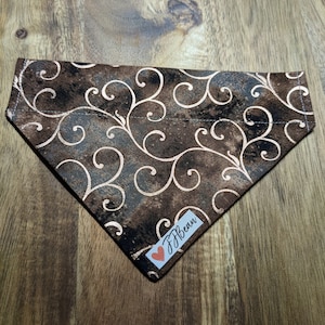 May include: A brown and tan dog bandana with a swirling, floral pattern. The bandana has a tag that reads "JBean" with a red heart. The fabric is a dark brown with lighter brown and tan accents.