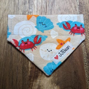 May include: A beige bandana with a sea-themed print featuring red crabs, blue seashells, and orange starfish. The bandana has a red heart with the text "JJBean" on it.