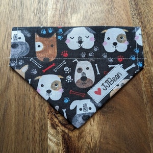 May include: A black dog bandana with a repeating pattern of cartoon dog faces, paw prints, and bones. The bandana has a white tag with the text "JJBean" and a red heart.