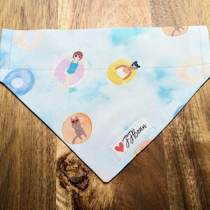 May include: A light blue dog bandana with a watercolor design of people and cats in inner tubes. The bandana has a white label with the text "JJ Bean" in black.