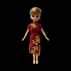 May include: A doll wearing a red crocheted dress with gold floral embroidery. The doll has blonde hair and blue eyes.