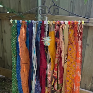 May include: A decorative metal hanger adorned with colorful beaded accents, suspending a collection of vibrant, patterned scarves. A dragonfly charm hangs from the center. The scarves display a variety of colors and designs, creating a bohemian aesthetic.
