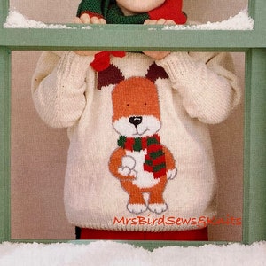 Może przedstawiać: Małe dziecko w białym swetrze z dzianiny z wzorem brązowego i pomarańczowego lisa. Lis nosi zielono-czerwoną szalik w paski. Dziecko patrzy przez okno z śniegiem na parapecie.