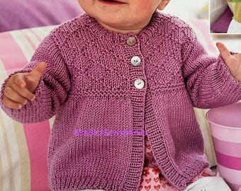 May include: A baby wearing a knitted, button-up cardigan in a shade of purple. The cardigan has a textured design on the upper chest and sleeves. The baby is also wearing white pants and a floral print shirt. A pink and white striped bucket is next to the baby.