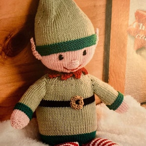 May include: A knitted elf doll with a green hat, red and white striped pants, and brown shoes. The elf is wearing a green sweater with a black belt and a brown button.