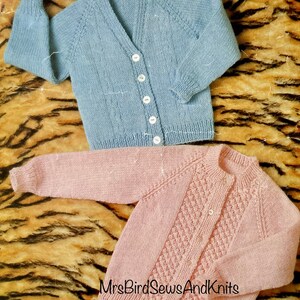 May include: Two knitted baby cardigans. One is light blue with a V-neck and white buttons. The other is pink with a round neck and white buttons. The cardigans are displayed on a tiger-striped background.