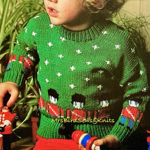 May include: A green knitted sweater with white floral accents and toy soldier designs on the sleeves and front. The sweater is worn by a child. The text "MrsBirdSews&Knits" is visible on the sweater.