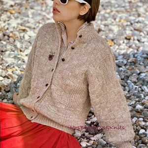 May include: A woman wearing a tan, chunky knit cardigan with a diamond pattern, button closure, and a small pin. She is wearing white-framed sunglasses and a red skirt. The image includes the text "CHUNKY" and measurements in inches and centimeters.