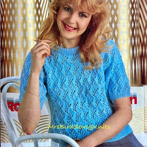 May include: A light blue knitted sweater with a lacy pattern. The sweater has short sleeves and a round neckline. The sweater is shown on a model wearing a grey skirt.