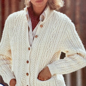 May include: A white cable knit cardigan with wooden buttons and pockets. The cardigan is worn over a white button-down shirt with a red patterned scarf.