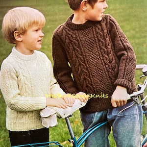 Op de afbeelding: Twee jongens in gebreide truien staan ​​naast een fiets. De jongen links draagt ​​een crèmekleurige trui. De jongen rechts draagt ​​een bruine kabelbreitrtrui.