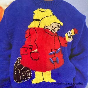 Könnte beinhalten: Ein blauer Strickpullover mit einer roten und gelben Comicfigur, die einen gelben Hut trägt und einen roten Eimer hält. Die Figur trägt eine braune Tasche mit den Buchstaben "PB" darauf. Der Pullover hat den Text "MrsBirdSewsgKnits" unten.