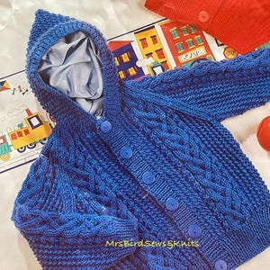 May include: A blue knitted baby cardigan with a hood and button closure. The cardigan features a cable knit pattern and blue buttons.