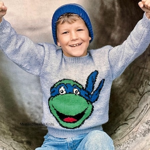 May include: A light blue long-sleeved knit sweater featuring a graphic of a green turtle character with a blue mask on the front. The sweater is paired with a matching blue knit beanie. The text MrsBirdSews&Knits is visible on the lower left side.