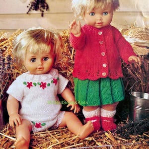 May include: Two dolls dressed in knitted outfits. One doll wears a white top and shorts with red floral embroidery. The other doll is in a red cardigan with heart-shaped buttons, a green skirt, and red and white socks.