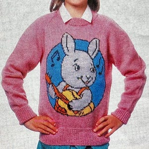 May include: A pink knitted sweater featuring a cartoon rabbit playing a guitar. The rabbit is set against a blue background with musical notes. The sweater has long sleeves and a crew neck, suitable for children.