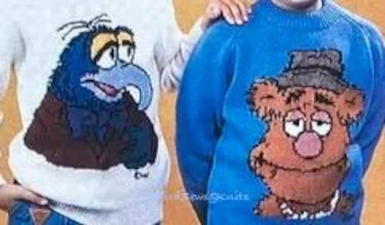 May include: Two knitted sweaters featuring characters from the Muppets. The sweater on the left is white with a blue and brown image of a blue bird with a bow tie. The sweater on the right is blue with a brown image of a bear wearing a hat.