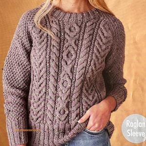 Puede incluir: Un suéter de punto trenzado de color malva con cuello redondo y mangas raglán. El suéter presenta intrincados patrones de trenzas en la parte delantera y en las mangas. La imagen muestra la textura y los detalles del diseño. Talla 81-127 cm.