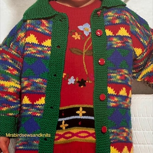 May include: A child's knitted cardigan in vibrant colours, featuring a green collar and red buttons, worn over a red sweater with floral embroidery. The cardigan has blue, yellow, and red geometric patterns. A knitted hat completes the outfit.