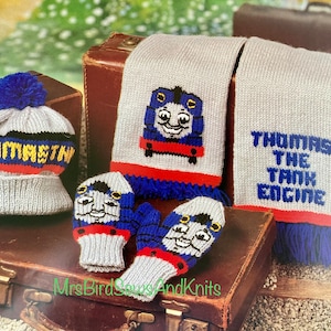 Puede incluir: Un conjunto de punto con Thomas la locomotora. El conjunto incluye un gorro, una bufanda y guantes. El gorro es gris con un pompón azul y las palabras "Thomas the Tank" en amarillo y rojo. La bufanda es gris con una raya azul y roja y una flecos azules. Los guantes son grises con una raya azul y roja y un puño azul. El conjunto se muestra sobre dos maletas vintage.