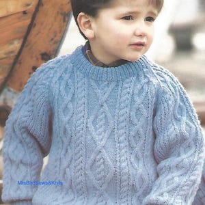 May include: A pale blue cable-knit jumper with a round neck. The jumper has a textured pattern of vertical and diamond-shaped cable designs. The sleeves are long with ribbed cuffs. The text "MrsBirdSews&Knits" is visible.