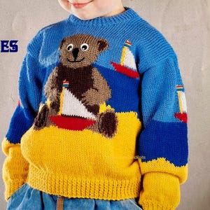 Può includere: Un maglione lavorato a maglia blu, giallo e marrone con un orsacchiotto e barche a vela. Il maglione ha uno sfondo blu con un fondo e polsini gialli. L'orsacchiotto è marrone con una barca a vela bianca e rossa. Le maniche hanno barche a vela.