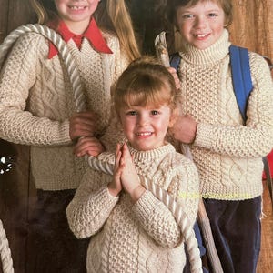 May include: Three children wearing cream-colored Aran knit sweaters. The sweaters have intricate cable and textured patterns. The image includes the text "Aran" and measurements of 20-32 inches. The children are smiling and holding a rope.