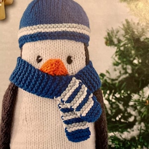 May include: A knitted penguin doll wearing a blue hat and scarf. The penguin has a white body, black wings, and orange feet and beak. The hat has a blue pom-pom and a white stripe. The scarf is blue with white stripes.