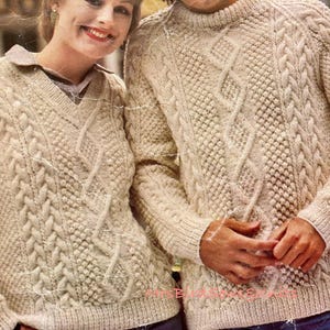 May include: Two cream-coloured cable knit jumpers with a textured pattern. The jumpers are worn by two people standing next to each other.