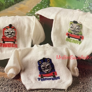 Könnte beinhalten: Drei weiße Strickpullover mit Thomas der Lokomotive-Charakteren. Die Pullover zeigen James, Henry und Thomas mit ihren Namen, die unter jedem Charakter gestickt sind.