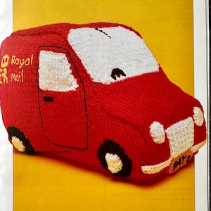 May include: A red knitted toy van featuring white windshields, headlights, and a front bumper. The side displays the yellow embroidered text Royal Mail with a crown symbol. The vehicle has black wheels with white centers and a small orange license plate.