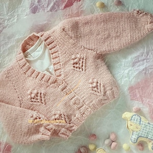 May include: A light pink, hand-knitted baby cardigan with leaf-shaped details. The cardigan is buttoned and has a ribbed collar and cuffs. A white undershirt is visible beneath the cardigan. The background includes a toy and decorative elements.