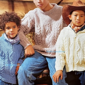 May include: Three children wearing cable-knit sweaters and jackets. One child wears a blue sweater, another a cream-colored zip-up jacket, and the third a light brown sweater. The child on the right wears a brown cowboy hat. All are wearing blue jeans.