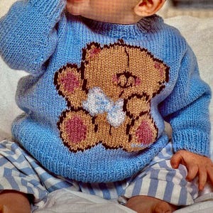 May include: A light blue knitted baby sweater featuring a brown teddy bear design with pink accents and a white bow. The sweater is part of a baby clothing set, paired with blue and white striped pants.