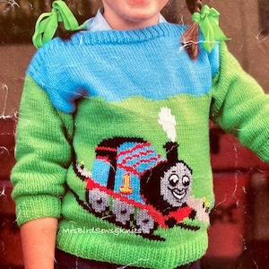 May include: A green, blue, and white knitted sweater with a Thomas the Tank Engine design. The sweater has a crew neck and long sleeves. The sweater is made of a soft, comfortable yarn.