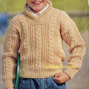 May include: A tan-colored, knitted sweater with a shawl collar and intricate cable knit detailing. The sweater has long sleeves and a ribbed hem and cuffs. The text "MrsBows&Knits" is visible on the lower right side of the sweater.