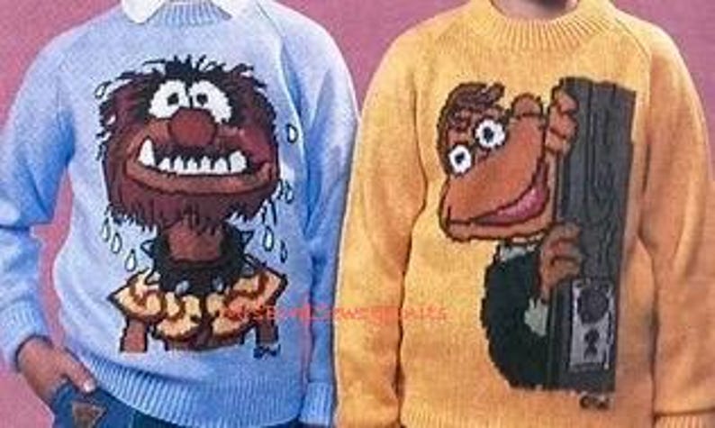 May include: Two knitted sweaters featuring characters from The Muppets. The sweater on the left is light blue with a brown and orange image of Animal, the drummer. The sweater on the right is yellow with a brown and orange image of Scooter, the Muppet who works in the control room.