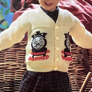 May include: A cream-colored, long-sleeved, V-neck knit cardigan featuring a button-down front with five white buttons. The sweater displays two identical black and grey train engine graphics with red accents and the word DONALD in red lettering.