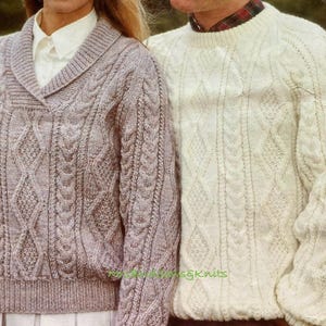 May include: Two cable-knit sweaters. One is a light gray with a shawl collar, the other is cream-colored with a crew neck. Both sweaters have intricate cable patterns and ribbed cuffs and hems. The sweaters are likely handmade.