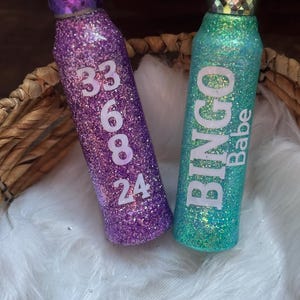 May include: Two glitter-covered water bottles. One is purple with the numbers 33, 6, 8, and 24 in white. The other is teal with the words "BINGO Babe" in white. Both bottles have decorative tops.