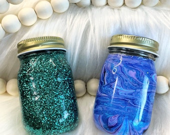 May include: Two small glass jars with lids. One jar is filled with teal glitter and the other jar has a blue and purple marbled design.