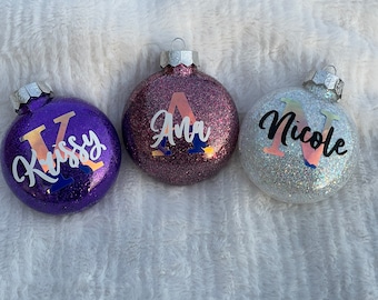 May include: Three glittery Christmas ornaments. One is purple with the name "Krissy", one is pink with the name "Ana", and the last is silver with the name "Nicole". Each ornament has a silver cap and a loop for hanging.