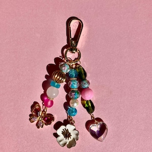 May include: A decorative keychain with a gold-colored clasp and a collection of colorful beads. The keychain features a gold butterfly charm, a white flower charm, and a clear heart-shaped charm. The beads are pink, blue, and gold.