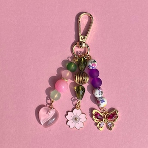 May include: A beaded keychain with a gold clasp. The keychain features a variety of beads in pink, green, purple, and gold, along with a pink heart, a pink flower, and a gold butterfly charm with red accents.
