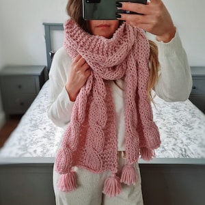 May include: A pink knitted scarf with a cable knit pattern and tassels. The scarf is draped around a person's neck and shoulders.