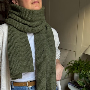 Peut inclure: Une écharpe en tricot vert olive est enroulée autour du cou. L'écharpe est épaisse et semble être faite d'un fil doux et texturé. L'écharpe est portée sur un t-shirt blanc et un jean bleu.