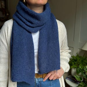 May include: A navy blue knitted scarf with a ribbed texture, worn around the neck. The scarf appears to be made of a soft material. The person is wearing a white top, a cream cardigan, and blue jeans. The scarf is a deep shade of blue.