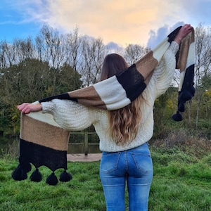 May include: A cream-colored sweater with a striped scarf in black, brown, and cream tones. The scarf has black tassels at the ends. The person is wearing blue jeans and black sneakers. The background includes a lake, trees, and a cloudy sky.